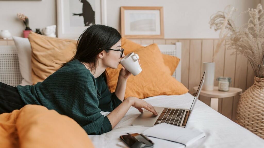 eLearning represented by a woman lying in bed drinking a beverage and looking at an open laptop