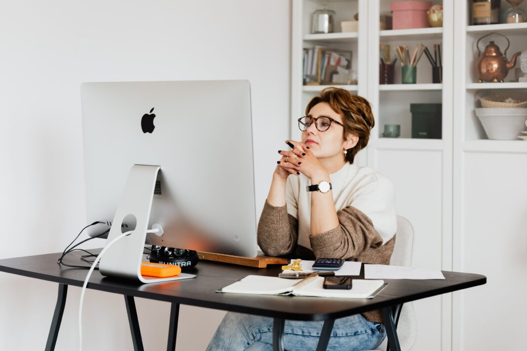 White woman with red hair sitting at white desk in front of computer with hands folded